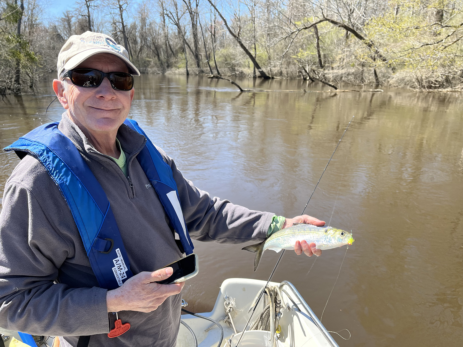 一名男子穿救生衣，站在河裡的船上，拿著一條閃亮的沙德魚。