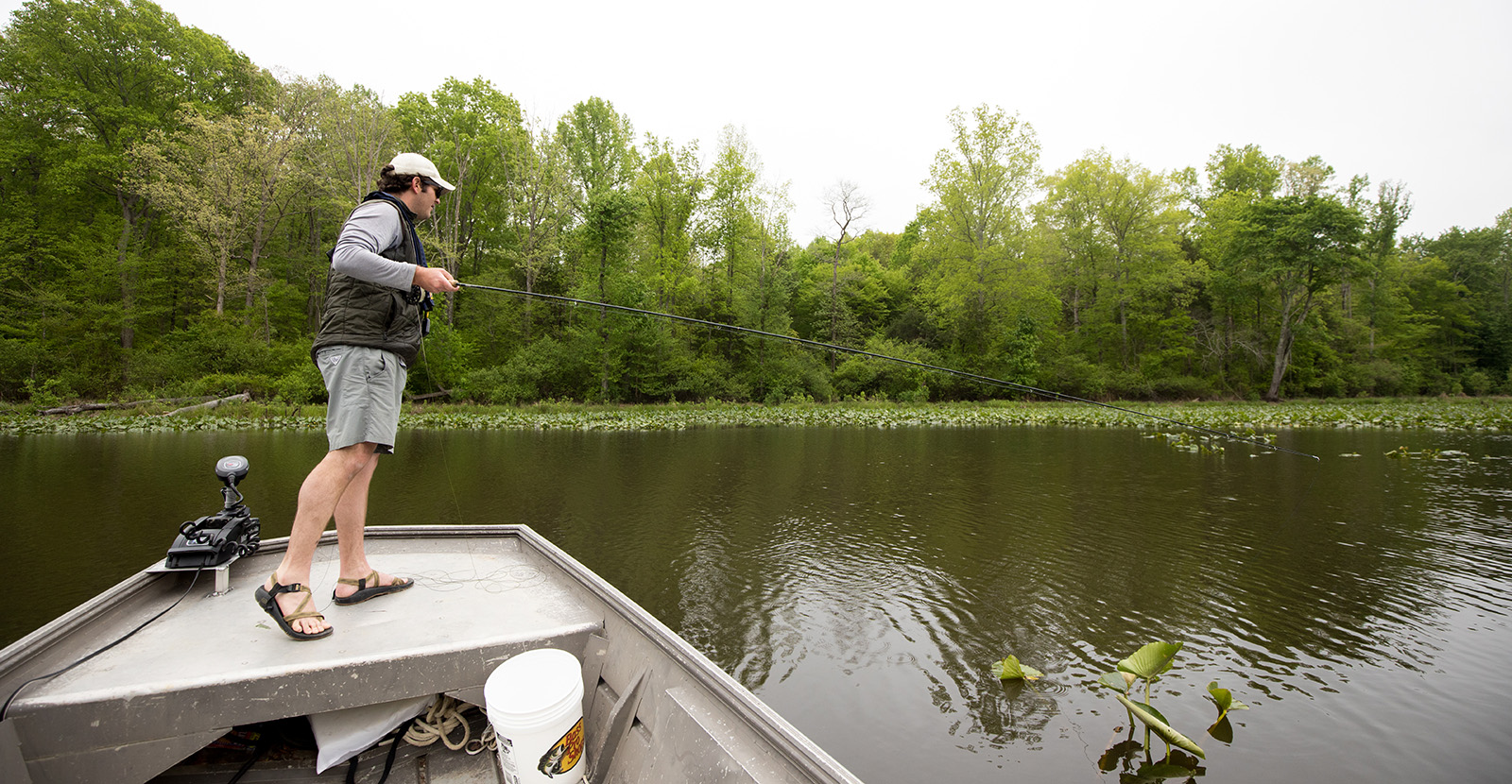 一個男子站在一艘船的弓邊,拿著釣竿,看著水中。