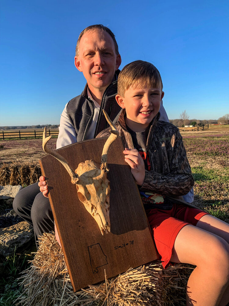 一個男人和他的太陽抱著一隻有框架的鹿頭骨的圖像,這是他們一起拍攝的第一隻鹿