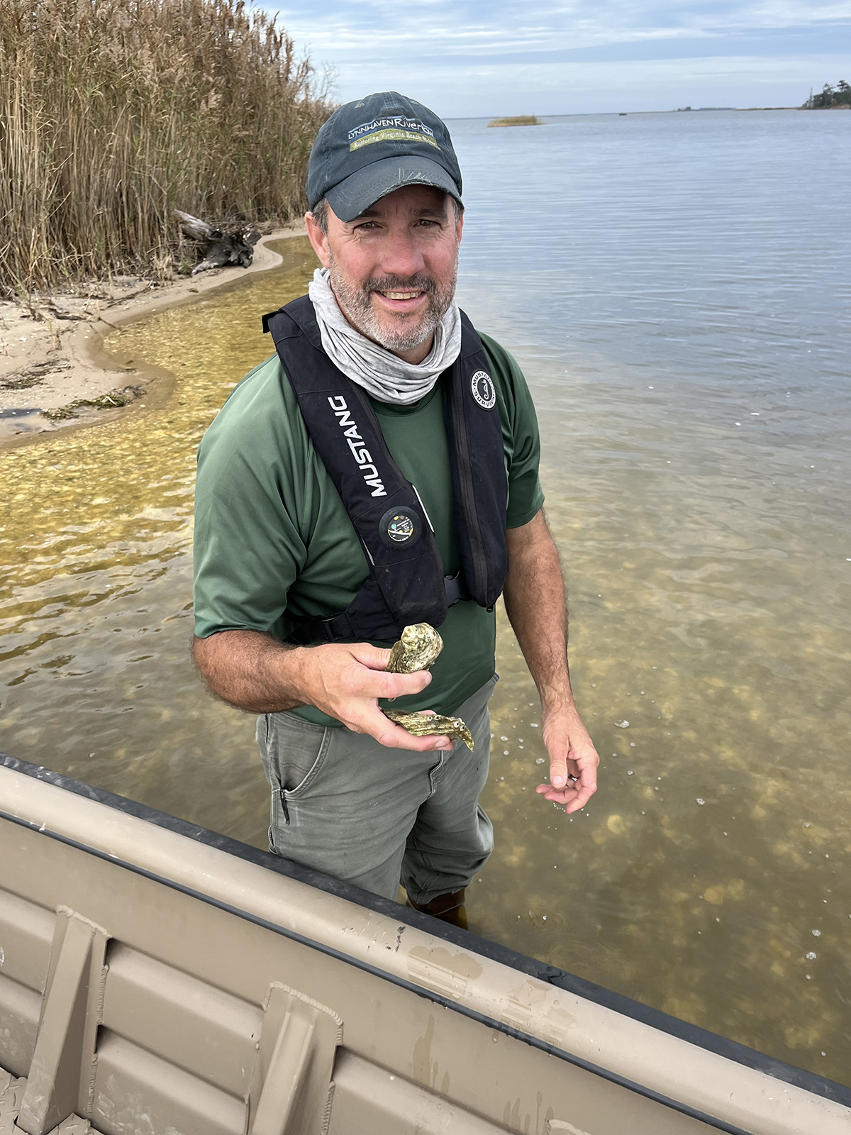 一位男子站在一艘船旁邊的水中,拿著牡蠣殼的照片。