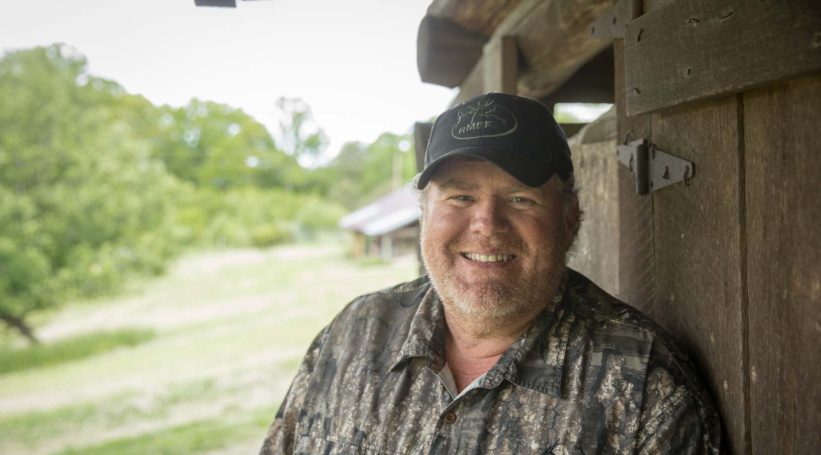 一張有鬍子的男人站在一棟粗糙的木建築旁邊的肖像照片。